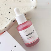 Medicube PDRN Salmon Peptide Skin Essence – Hydrating Serum in a pink glass bottle on a speckled surface.